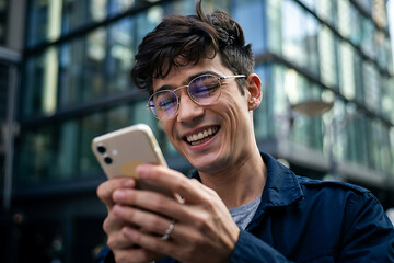 Young man laughing and using mobile phone outdoors with modern building backdrop capturing a moment of joy and connection in the digital age