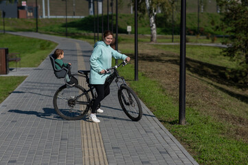 Caucasian woman riding bicycle with toddler son sitting behind her in child seat in park. 