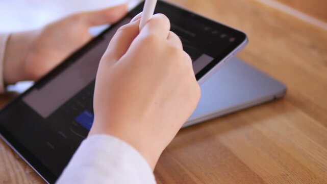Person is writing on a tablet with a pen. The tablet is on a wooden table. The person is using a stylus to write
