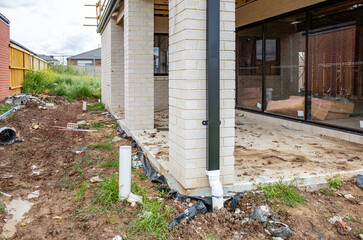 A newly built house in a suburban neighborhood with unfinished landscaping and debris. Exposed plumbing, downpipes, and raw soil around the exterior of a residential construction site in Australia