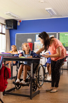 Asian teacher assists a girl in a wheelchair at a school classroom desk