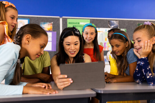 Asian teacher shows a tablet to a group of attentive children in a school classroom