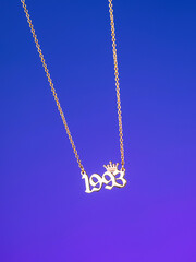 Close-up of a metallic gold pendant in the shape of "1993" on a chain, on a  purple background