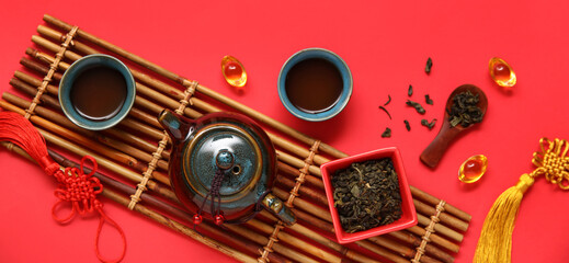 Bamboo mat with teapot, cups of tea, bowl and spoon with dry leaves on red background