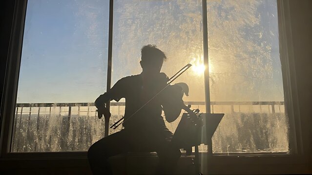 Silhouette of a violinist framed in golden light, performing as daylight fades behind a large window.