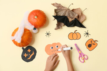 Child with supplies and decorations coloring Halloween paper pumpkins on yellow background