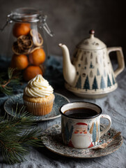 Cupcake with cream icing and a Christmas mug on a table with festive decoration; teapot, dried oranges, and pine sprigs.

