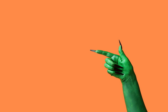 Green hand of witch pointing at something on orange background. Halloween celebration
