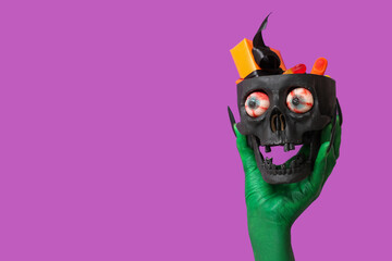 Green hand of witch holding skull with candies and gift box on purple background. Halloween celebration