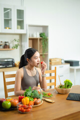 Asian woman holding green juice and apple, surrounded by fresh vegetables and salad in modern kitchen. Promotes healthy eating, fitness, and wellness lifestyle.