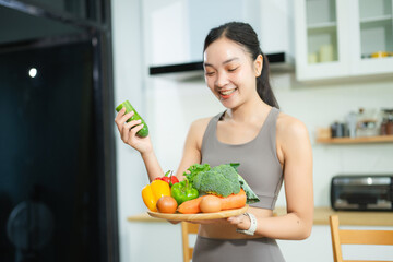 Smiling fit Asian woman in sporty wear showing tray of fresh vegetables and eggs in kitchen. Promotes healthy eating, clean food, wellness,