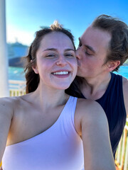 A man kisses his girlfriend on the cheek while she takes a selfie on the ocean coastline.
