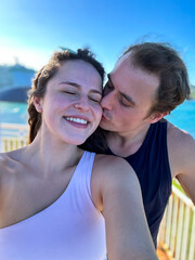 A man kisses his girlfriend on the cheek while she takes a selfie on the ocean coastline.