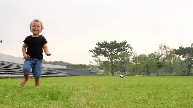 Playful toddler boy runs up to soccer ball on field and kicks it forward. Laughing, he turns and scampers in opposite direction, enjoying his mischief. Barefoot boy revels in carefree fun