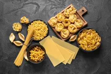 Cutting board and bowls with different types of raw pasta on black grunge background