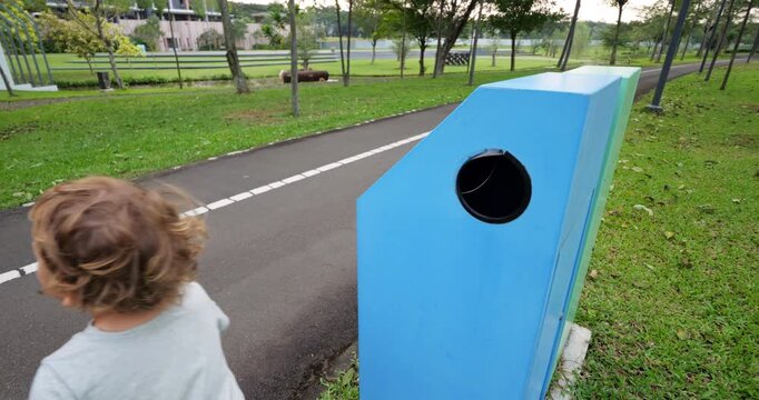 Toddler boy playfully runs to recycling bin and tosses empty plastic bottle into designated slot. He quickly runs back to get another bottle, joyfully engaging in eco-friendly task.