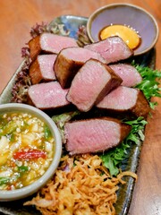 Medium-rare steak slices served with crispy onions, leafy greens, and two dipping sauces at a fine steak restaurant in Bangkok, Thailand.