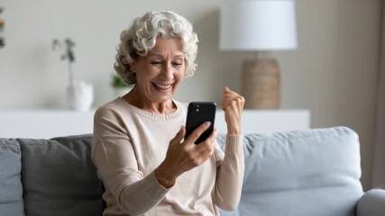 Excited Senior Woman Holding Phone in Gray Couch Celebrating Success Indoors in Beige Sweater with Cheerful Expression