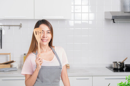 Beautiful Asian young woman joyfully poses in her white kitchen, holding cooking tool is wood spatula representing love for homemade meal.