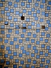 Fototapeta premium A geometric blue and white tiled wall features an intricate rectangular pattern, though several tiles are cracked or missing, revealing rusted metal and dark gaps beneath. 