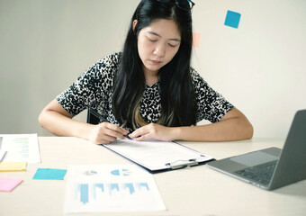 Young Asian business woman sitting at the table holding a pen, recording investments and planning...