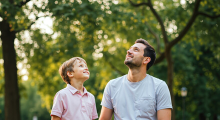 Father and son looking up together in sunny park. Light blue t-shirt, pink polo shirt, lush green trees. Summer family curiosity bonding concept