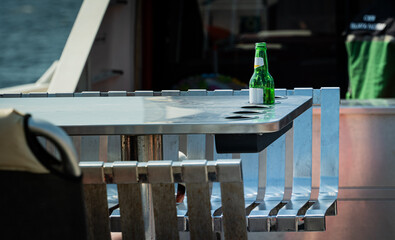 A green glass beer bottle with a white label sits on a stainless steel table with built-in cupholders on a boat, suggesting relaxation.