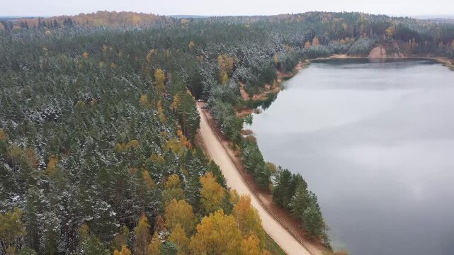 A drone glides over Ogres Zilie Kalni in Latvia, capturing a road tracing the quarry&rsquo;s edge before disappearing into snowy autumn woods filled with colorful, frosted trees.