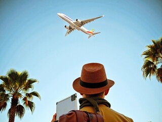 Man with Laptop Watching Airplane Fly Overhead Under Palm Trees