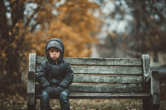 Young boy sitting alone on park bench during autumn, looking worried or sad, dressed warmly with hat and gloves, surrounded by fall foliage and trees in background