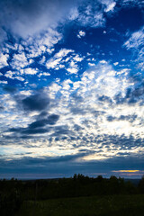 Obraz premium A dramatic sky unfolds during sunset, painted with swirling clouds in hues of blue, orange, and yellow. Trees silhouette the horizon, grounding the ethereal scene in nature’s quiet rhythm.