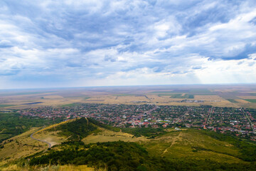 Obraz premium A small town nestles between forested hills and patchwork fields under a partly cloudy sky, blending rural life with nature in a serene, panoramic landscape.