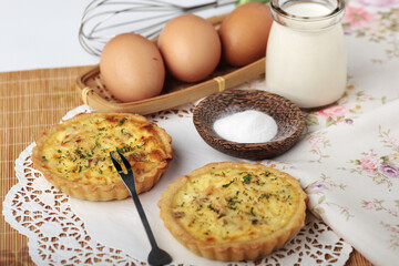 Two savory mini quiches are displayed on a white doily next to ingredients like eggs, milk, and salt, with a wire whisk in the background