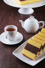 A tea set with a white teapot and a cup of tea is arranged alongside a platter of sliced Lapis Surabaya cake on a dark wooden table.
