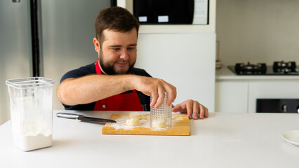 A man in a red apron flattens dough pieces using a glass on a floured cutting board in a modern kitchen, preparing homemade baked goods with focus and care.