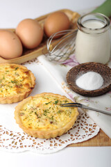 Two savory mini quiches are displayed on a white doily next to ingredients like eggs, milk, and salt, with a wire whisk in the background