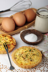 Two savory mini quiches are displayed on a white doily next to ingredients like eggs, milk, and...