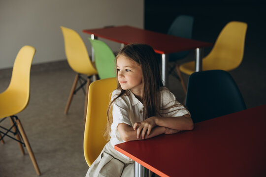 a 7-year-old girl sitting at a table with colored chairs in a large room, classroom or school cafeteria - Powered by Adobe