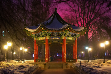 Fototapeta premium Colorful Chinese pavilion covered in snow, illuminated by lanterns in a city park at dusk, winter evening, Chengdu, China. Peaceful atmosphere.