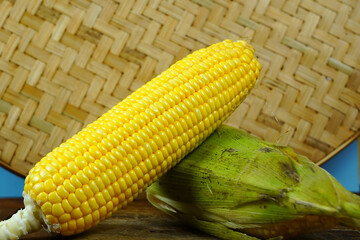 Two fresh, yellow corns, one partially husked, rest on a rustic wooden cutting board with a woven basket in the background.
