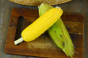 Two fresh, yellow corns, one partially husked, rest on a rustic wooden cutting board with a woven basket in the background.