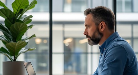 Man in blue shirt gazes thoughtfully out a window next to a potted plant