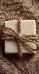 Square bar of light beige soap, tied with a natural twine bow, on a textured brown burlap fabric