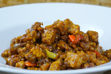 A white plate of Indonesian stir-fried tempeh with soy sauce, chili, and other spices.