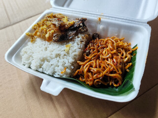 Styrofoam box containing mixed rice, with white rice, fried salted fish, and fried noodles covered in banana leaves, ready to eat.