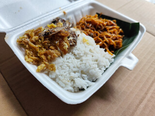 Styrofoam box containing mixed rice, with white rice, fried salted fish, and fried noodles covered in banana leaves, ready to eat.