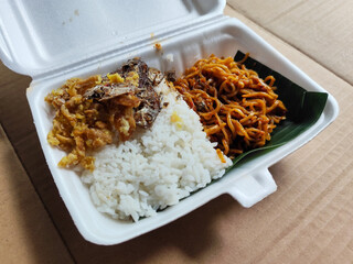 Styrofoam box containing mixed rice, with white rice, fried salted fish, and fried noodles covered in banana leaves, ready to eat.