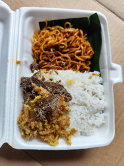 Styrofoam box containing mixed rice, with white rice, fried salted fish, and fried noodles covered in banana leaves, ready to eat.