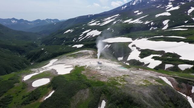 Aerial view of a majestic geyser releasing a plume amidst snow-capped mountains and green valleys, contrasting textures and tones, Kamchatka Krai, Russia.