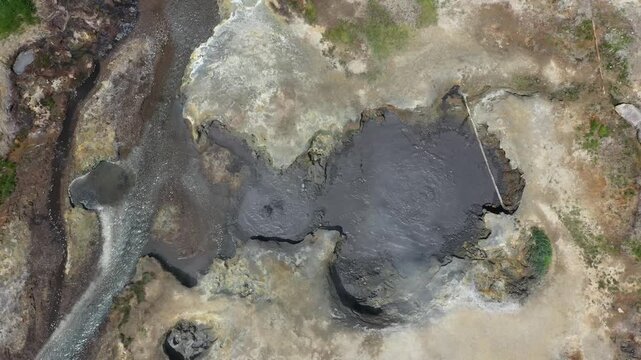 Aerial view of bubbling mud volcano contrasting with a winding stream and geothermal terrain, creating a textured mosaic of earth tones, Kamchatka Krai, Russia.